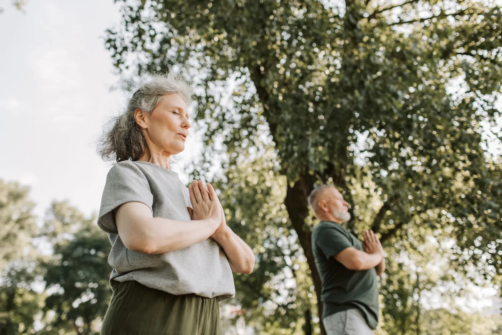 A woman with gray hair stands with her eyes closed and hands pressed together in a prayer pose, with a blurred man in the background doing the same under green trees.