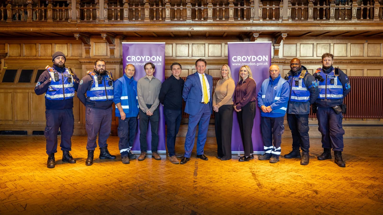 A group of eleven adults, some in safety vests and uniforms, and others in smart casual attire, stand in a line in front of two purple banners that say "CROYDON".