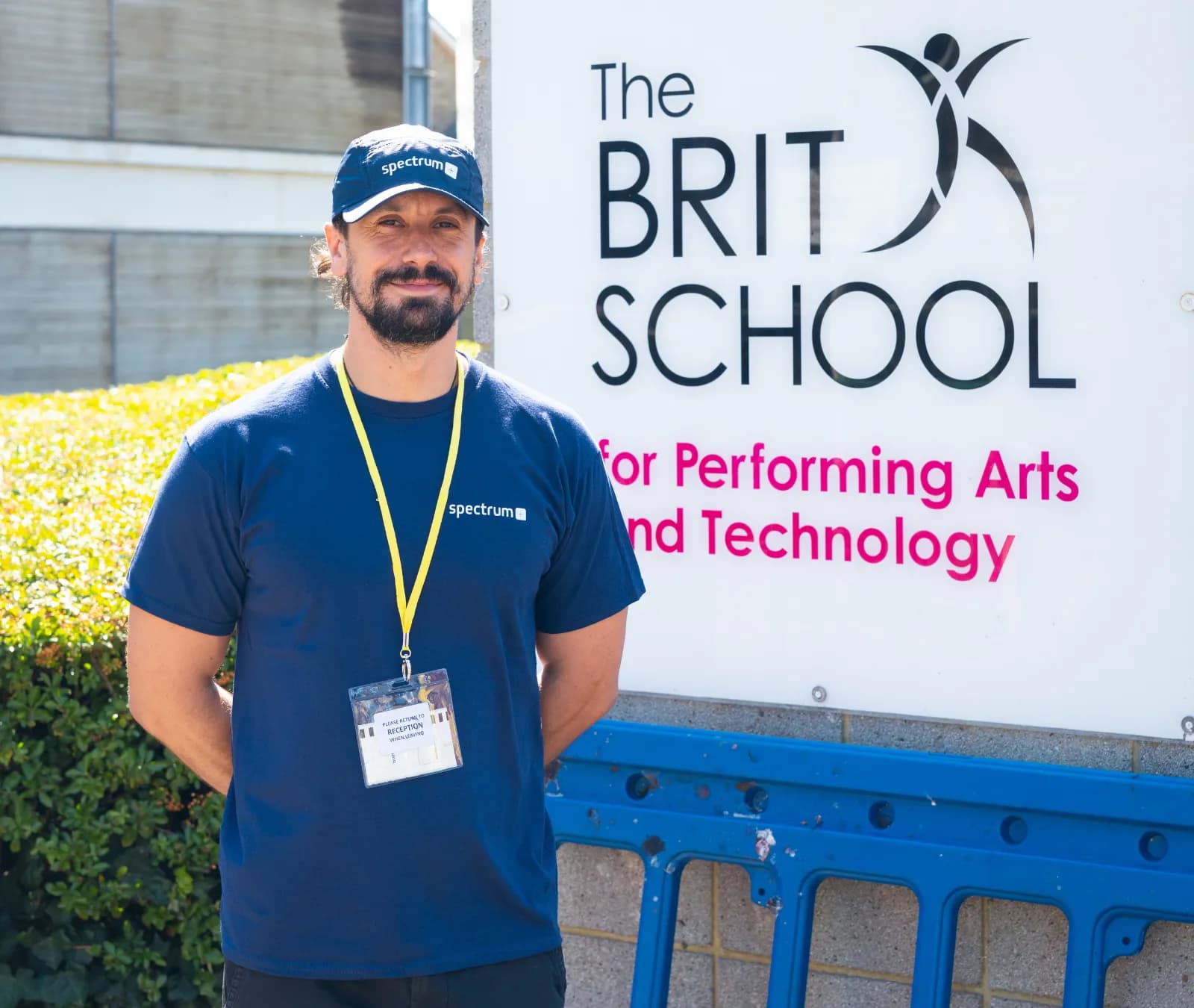 A man wearing a dark blue t-shirt and baseball cap with a Spectrum logo, and a yellow lanyard with an ID badge, stands smiling in front of a white sign that reads 'The BRIT SCHOOL for Performing Arts and Technology'.