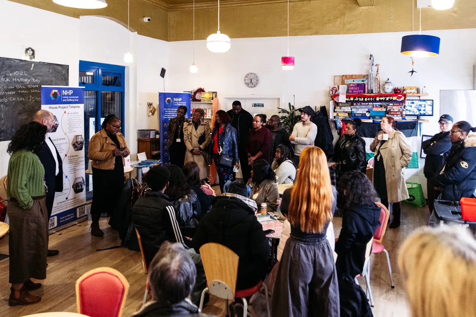 A group of people are gathered indoors, some standing and some seated, listening to a woman speaking in the foreground of a large room with white walls, blue doors, and hanging lamps.